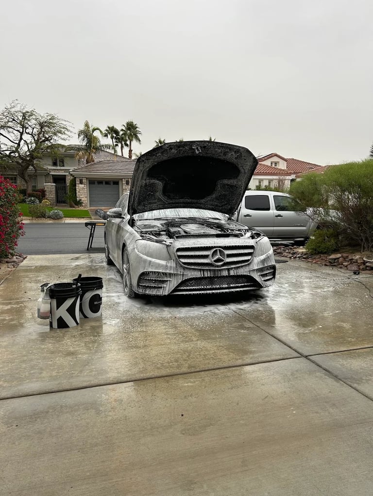 White Mercedes with open hood on driveway, being washed in suburban setting with palm trees and homes in background
