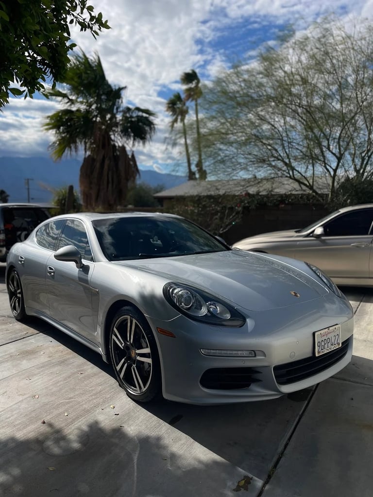 Silver Porsche Panamera parked on a paved surface with palm trees and mountains visible in the background under a partly cloudy sky
